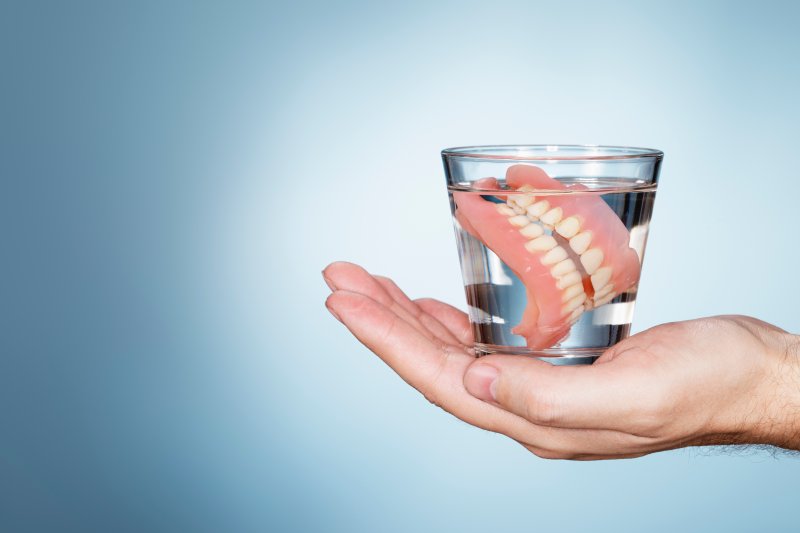 Hand holding a glass of water with dentures floating in it over blue background